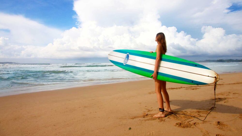 Surfer girl standing on beach with board looking at ocean